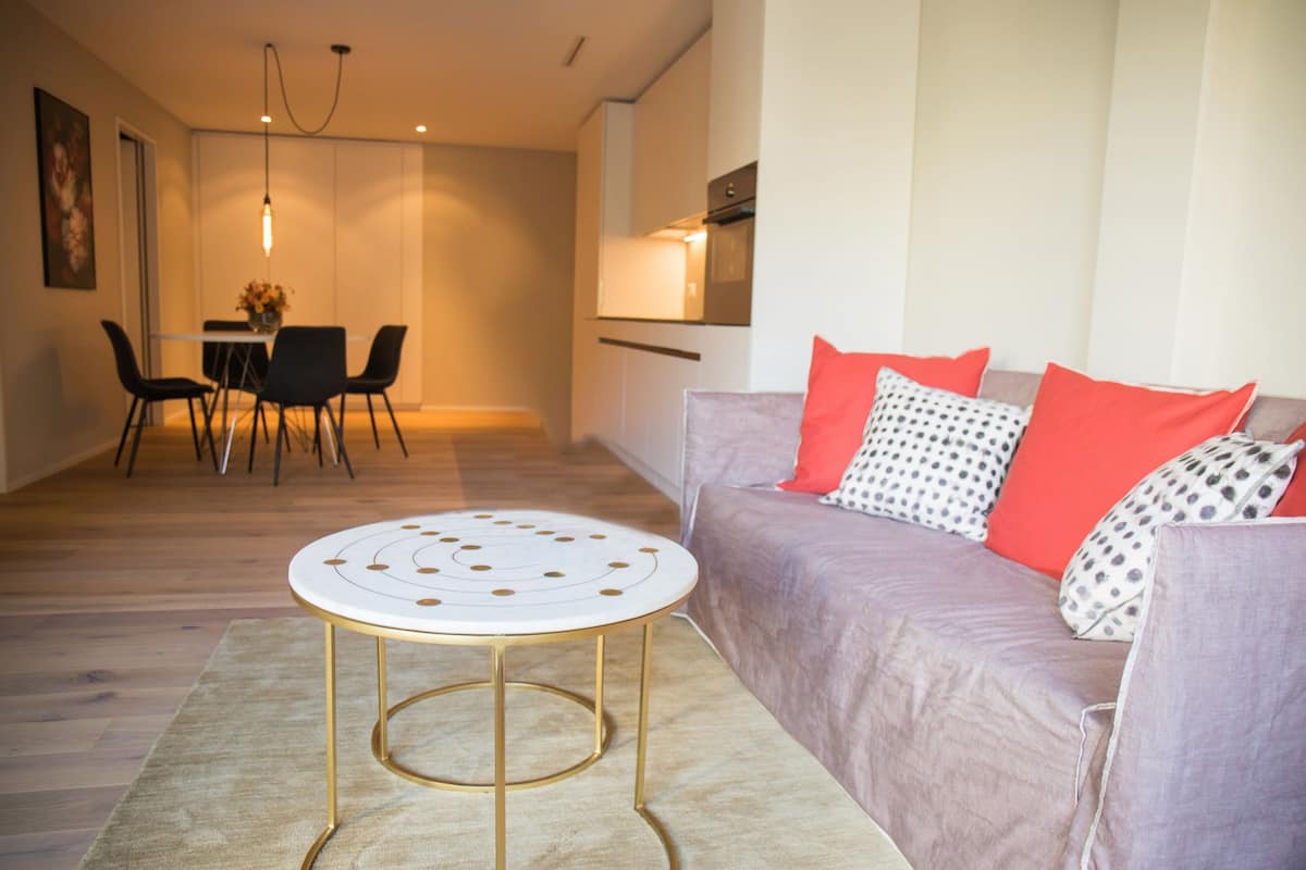 Una sala de estar moderna con una mesa redonda, un sofá con cojines estampados y un comedor con sillas y un jarrón de flores.
