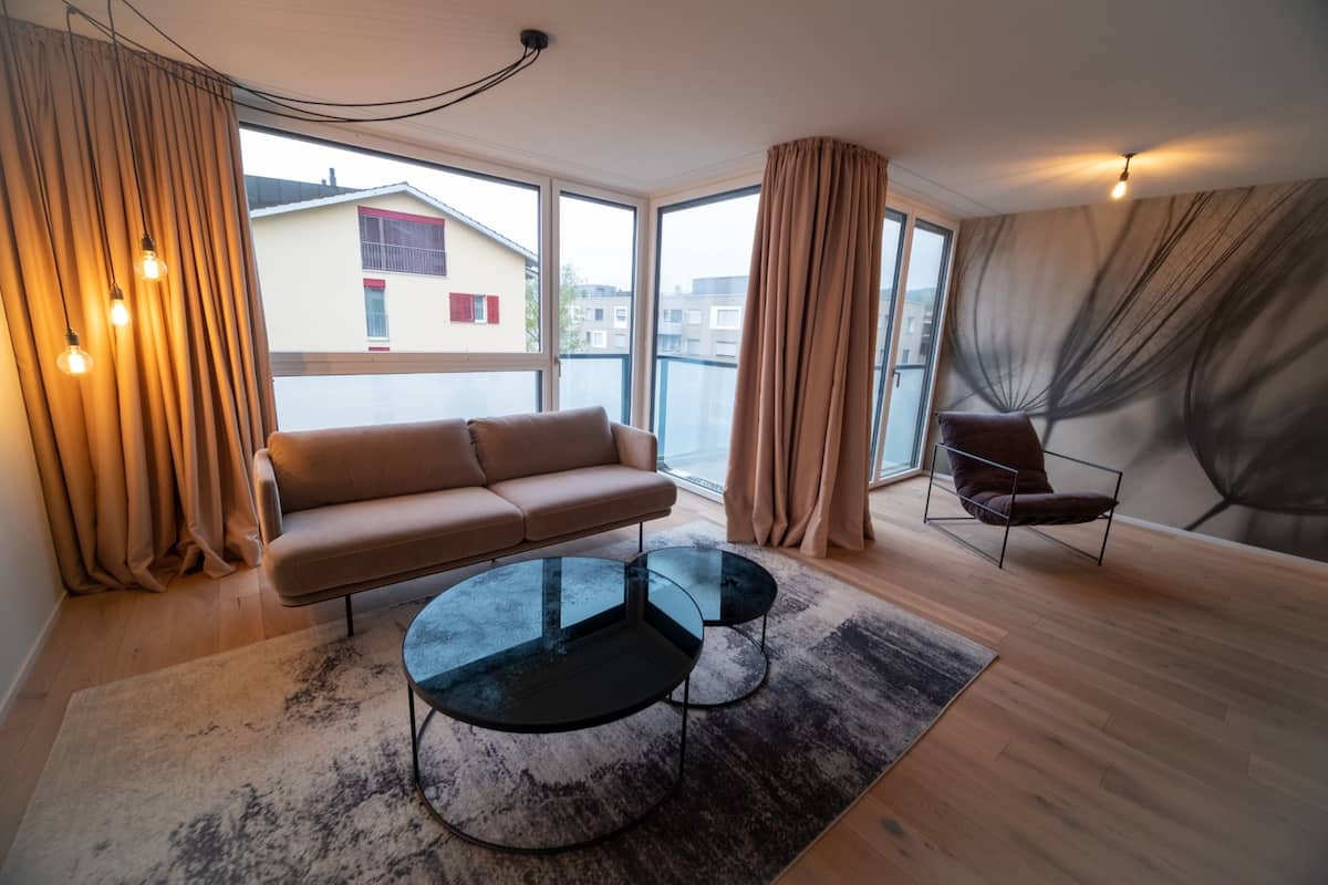 Un salón moderno con un sofá, dos mesas redondas y una silla. Amplias ventanas ofrecen vistas a edificios exteriores.