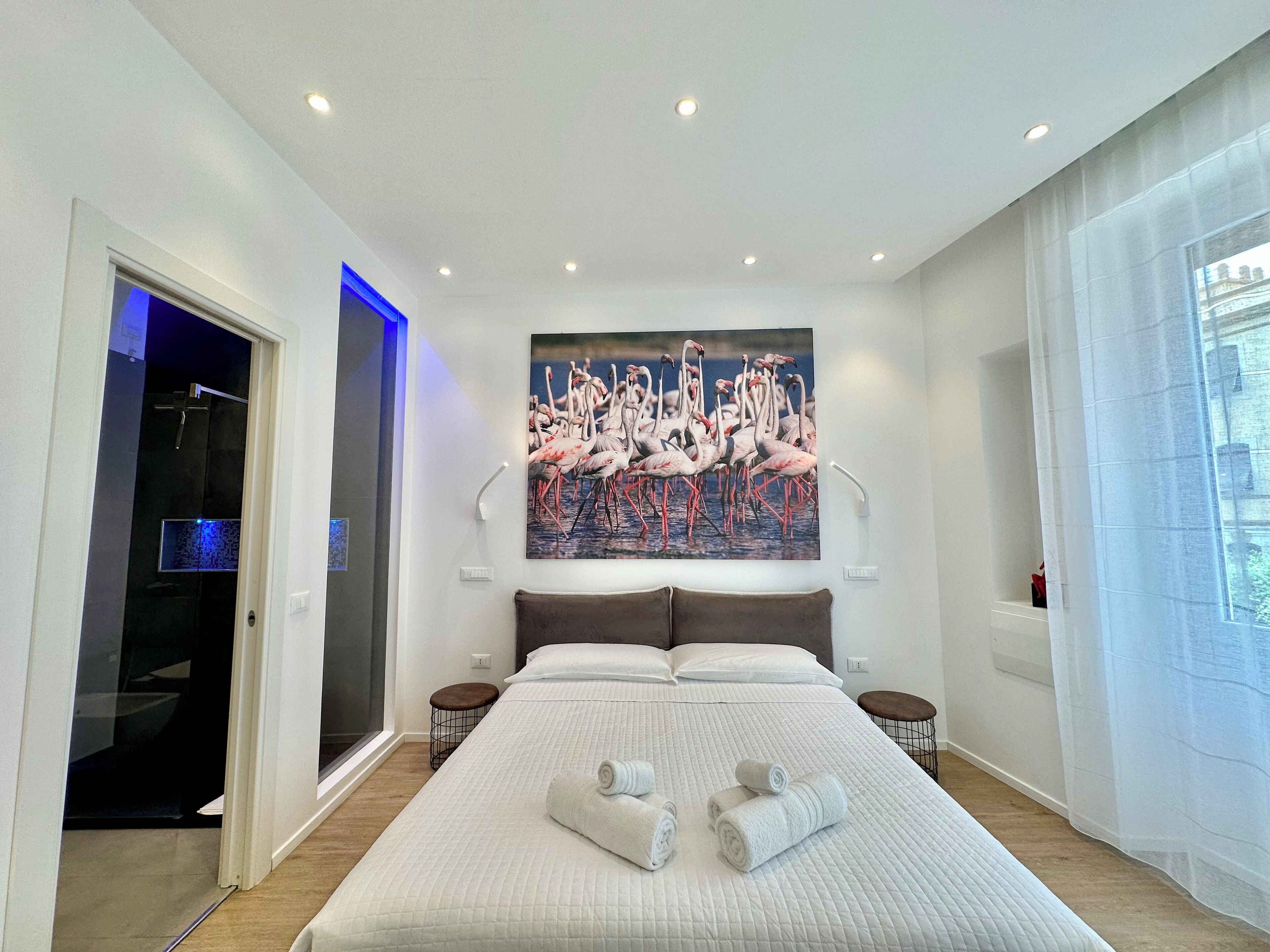 Een moderne hotelkamer met een groot bed, een flamingo schilderij, een inloopkast en een badkamer met een glazen deur.