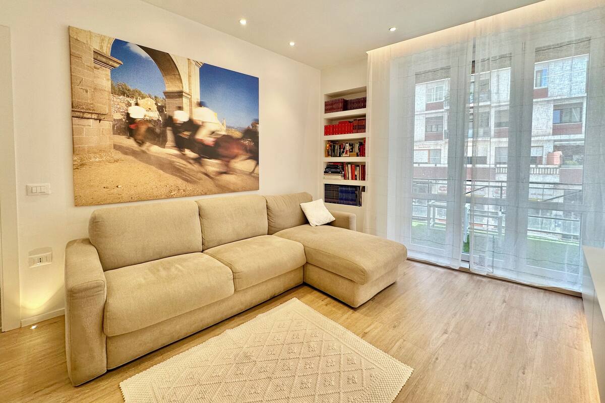 A modern living room with a beige sectional sofa, a large abstract painting, a bookshelf, and a view through a large window.