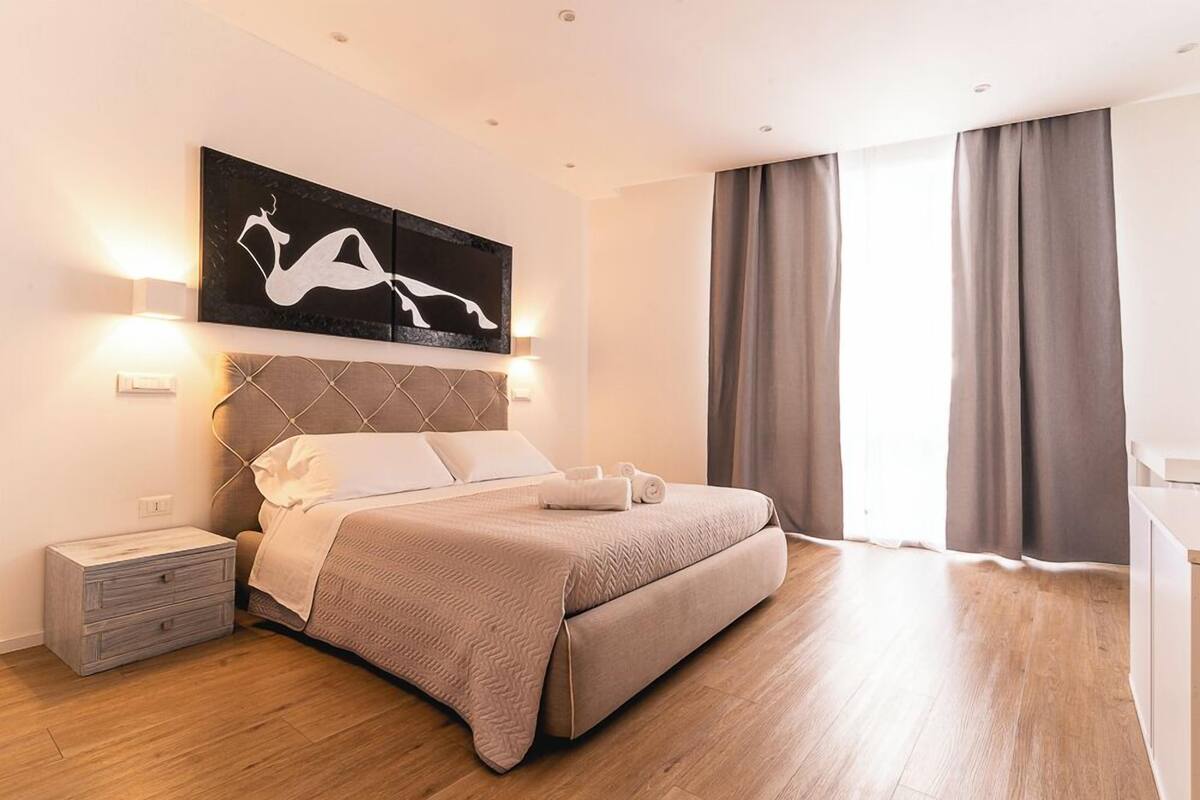 A modern bedroom with a large bed, a nightstand, a wall-mounted artwork, and a window with curtains.