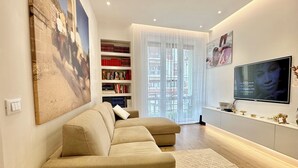 A modern living room with a beige sofa, a large painting, a bookshelf, and a flat-screen TV. - J&O' Luxury Suite (Cagliari)