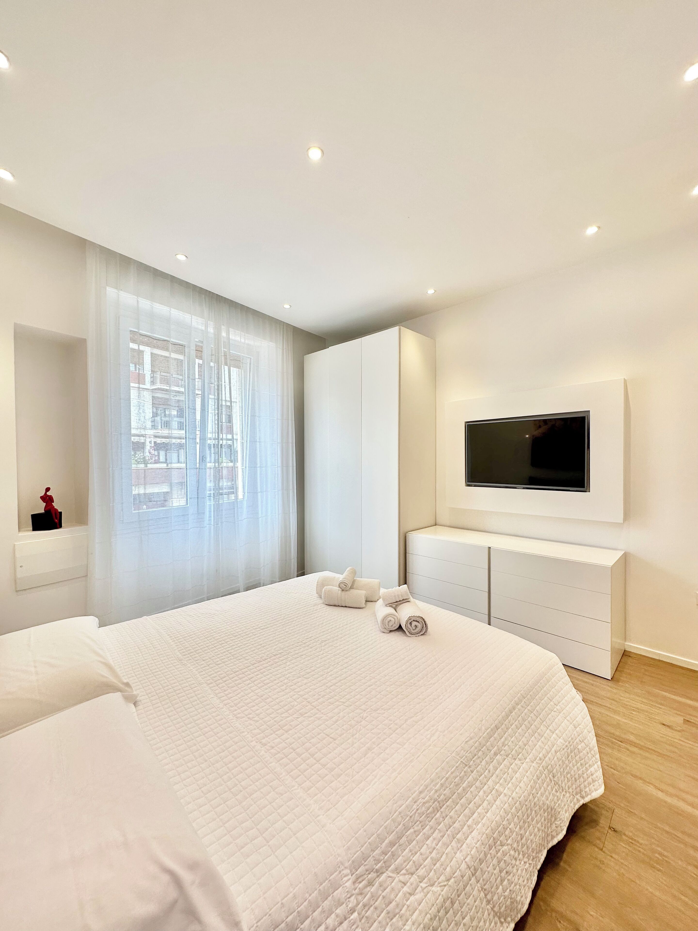 A modern bedroom with a large bed, a TV mounted on the wall, and a window with sheer curtains.