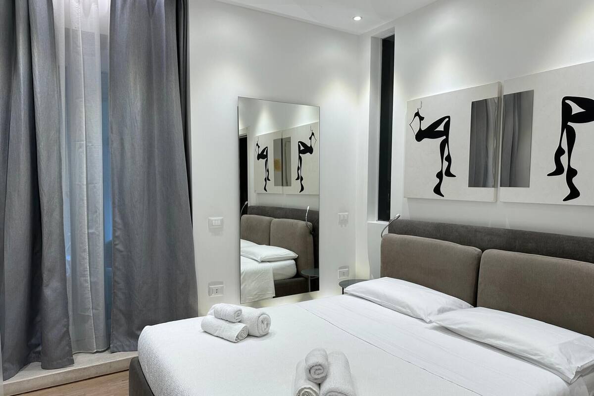 A modern hotel room with a large bed, grey curtains, a mirror, and abstract art on the walls.