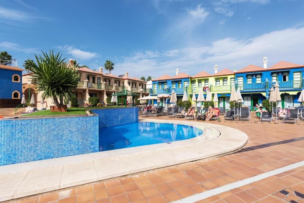 Una piscina con borde de azulejos, rodeada de sillones de descanso y sombrillas, con edificios coloridos al fondo.