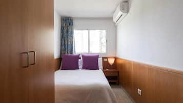 Un dormitorio con una cama, dos almohadas moradas, una mesita de noche, una lámpara y una ventana con cortinas.
