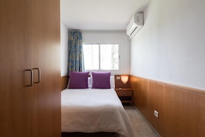 A bedroom with a bed, two purple pillows, a nightstand, a lamp, and a window with curtains.