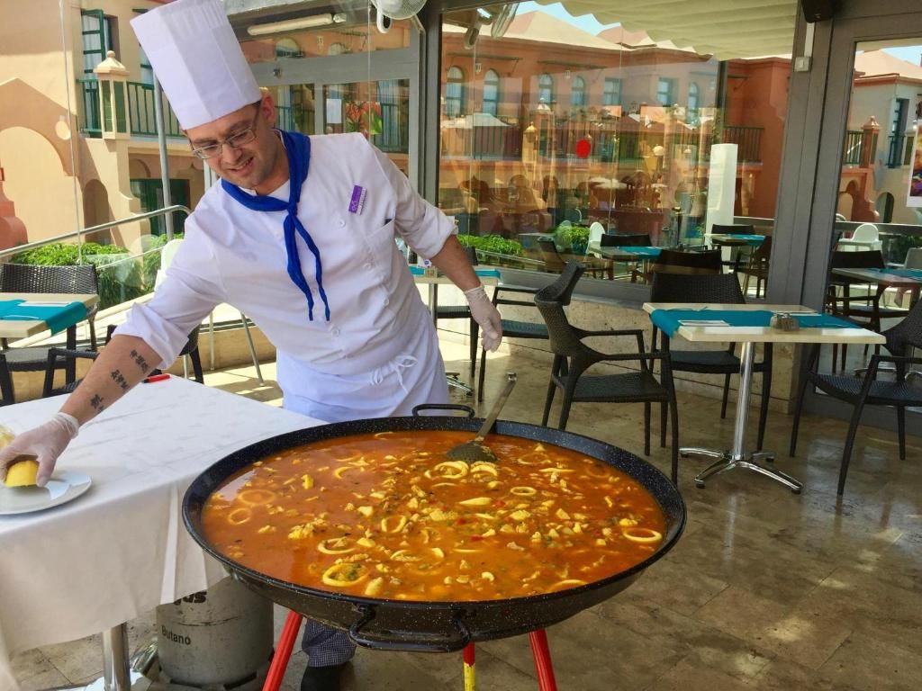A chef preparing a large pot of soup outdoors with tables and chairs in the background.