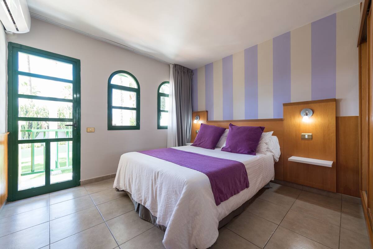 A bedroom with a bed, a nightstand, a wall-mounted light, and a window with curtains.