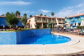 Un área de piscina con borde enlosado, sillas de descanso, sombrillas y palmeras.