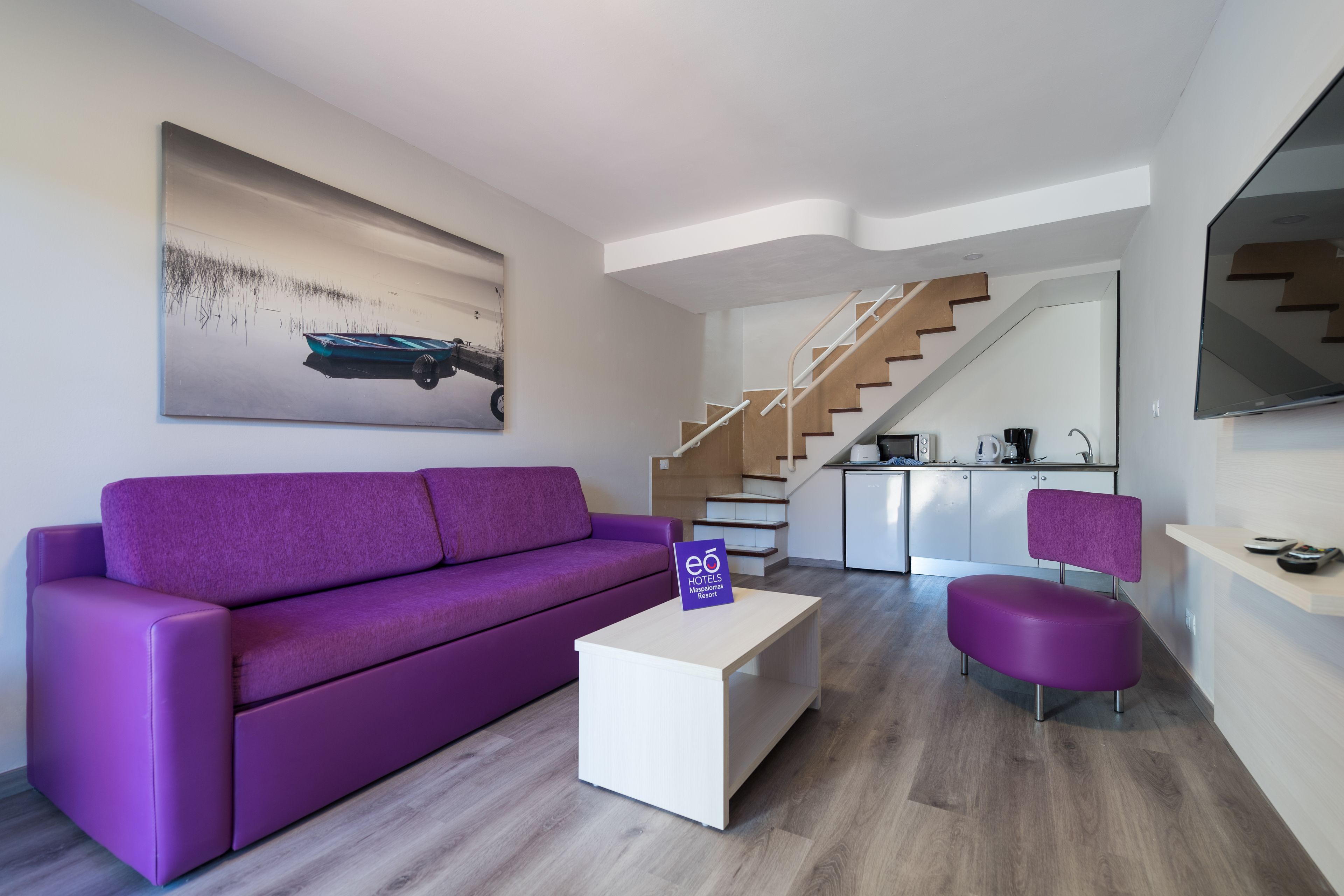 Un salón moderno con un sofá púrpura, una mesa de centro blanca y un cuadro de un coche en la pared. Una escalera conduce a una zona de cocina.