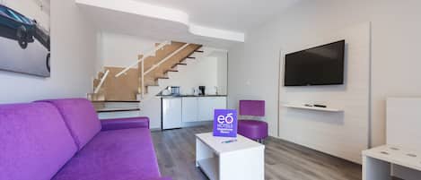 Una sala de estar moderna con un sofá púrpura, una mesa de centro blanca con un letrero, un televisor de pantalla plana y una escalera que lleva a una cocina.