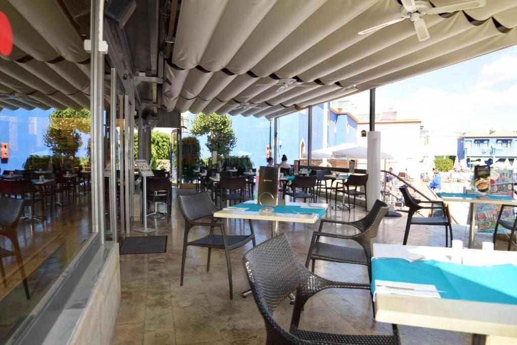 An outdoor cafe with tables and chairs, a ceiling fan, and a view of the sea.