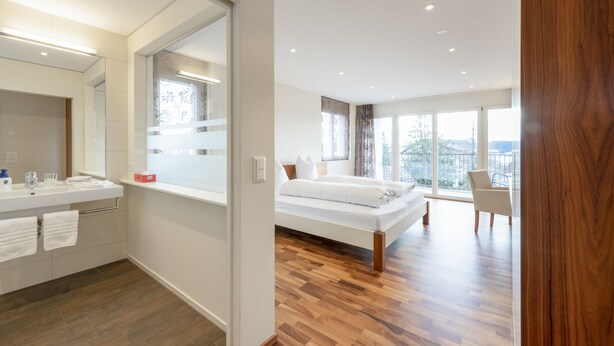 A modern bedroom with a large bed, a bathroom with a mirror and sink, and a view of the outdoors. - Hotel Rössli (Zuzwil)