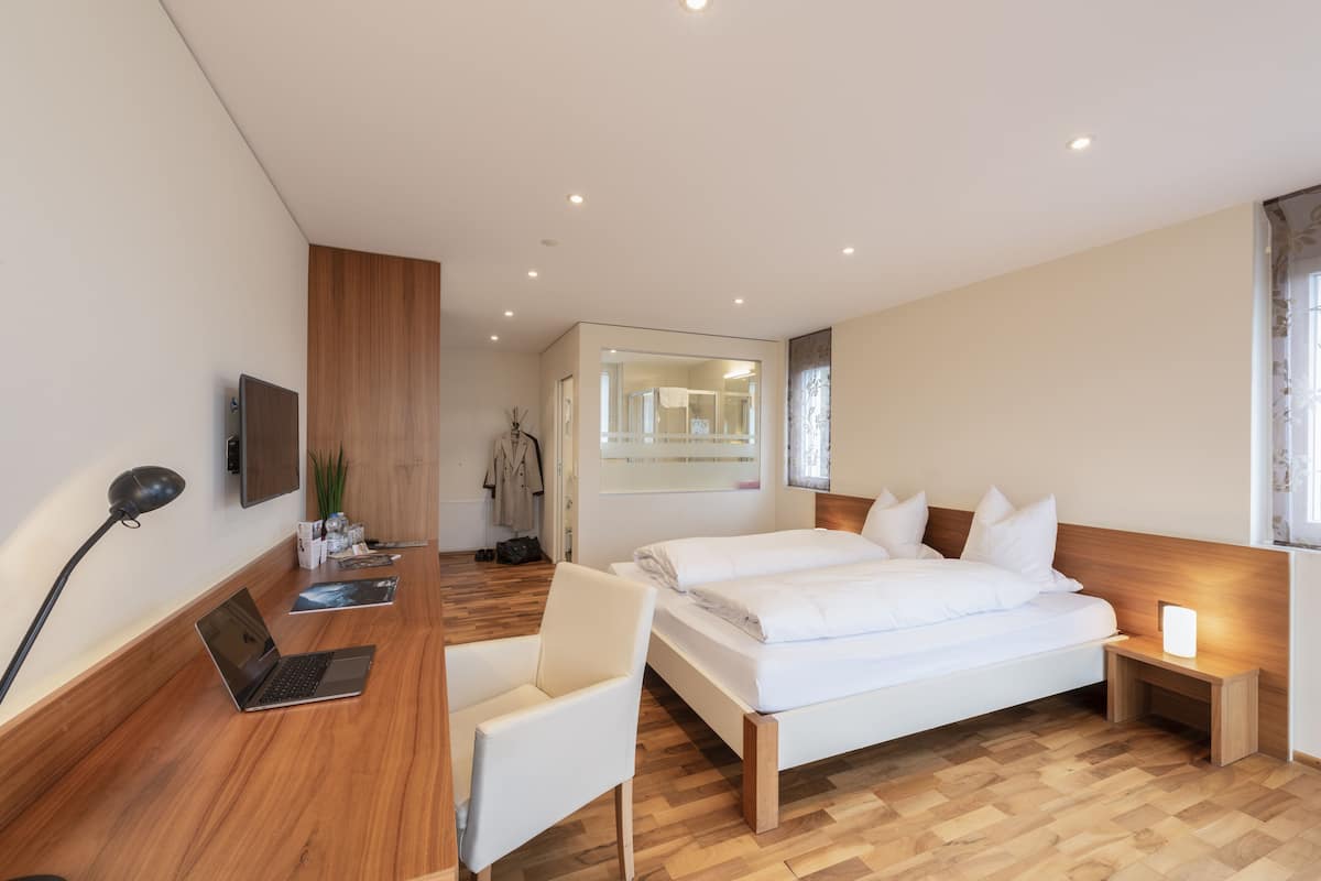 A modern hotel room with a wooden floor, two single beds, a desk with a laptop, a chair, and a bedside table with a lamp.