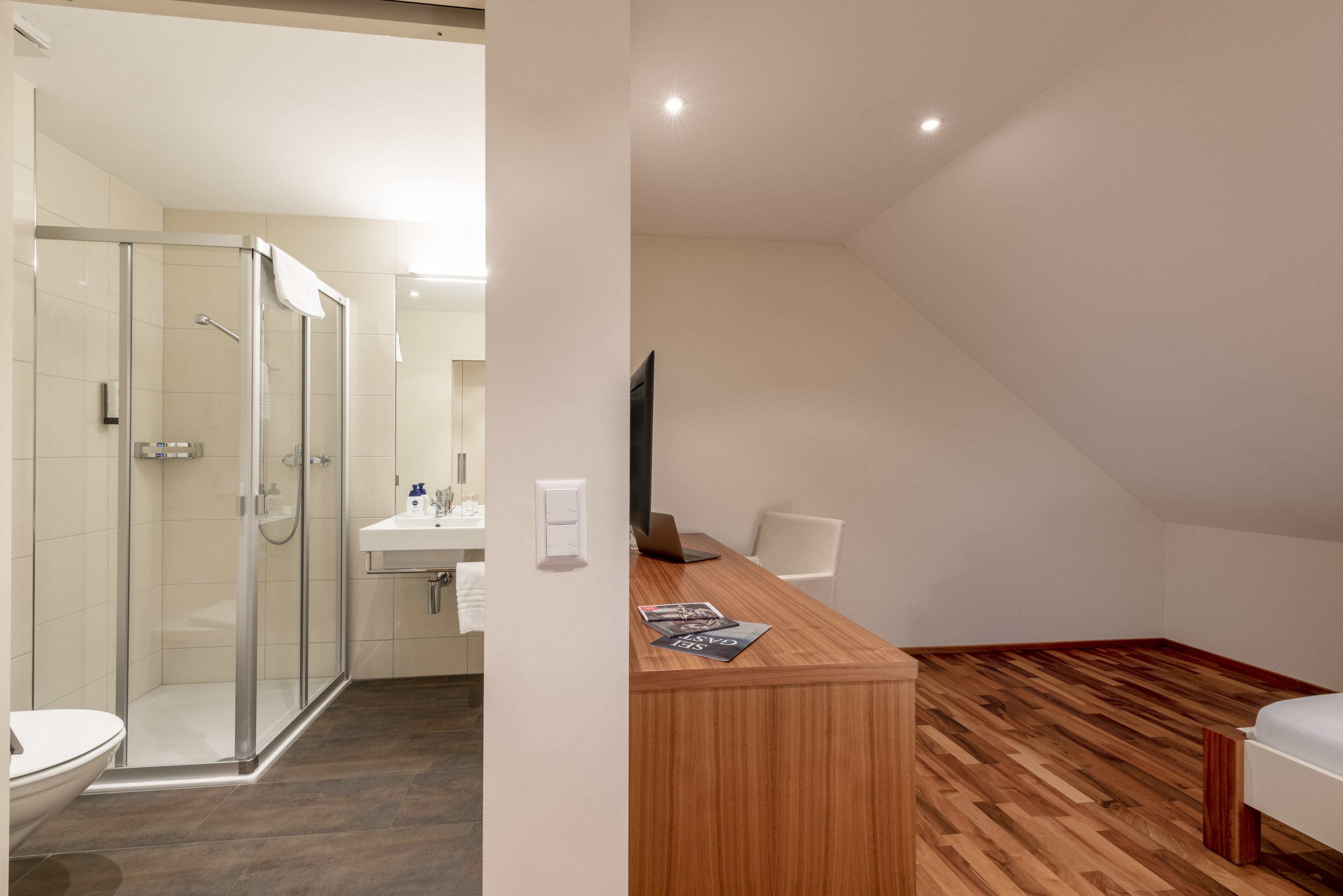 A modern bathroom with a glass shower enclosure, a white sink, and a wooden vanity with a laptop and magazine.