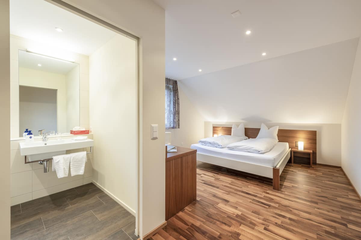 A modern bedroom with a wooden floor, a bed with white linens, a bedside table with a lamp, and a bathroom with a sink and mirror.