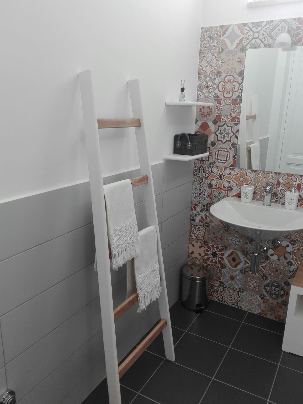 A bathroom with a white sink, patterned tiles, a white towel on a wooden ladder, and a small trash bin.