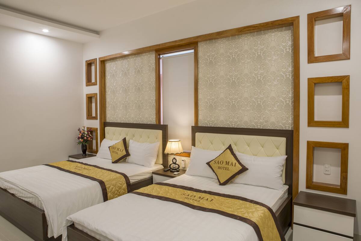 A hotel room with two beds, each with a headboard and a pillow with the hotel name. There is a nightstand with a lamp and a vase of flowers.