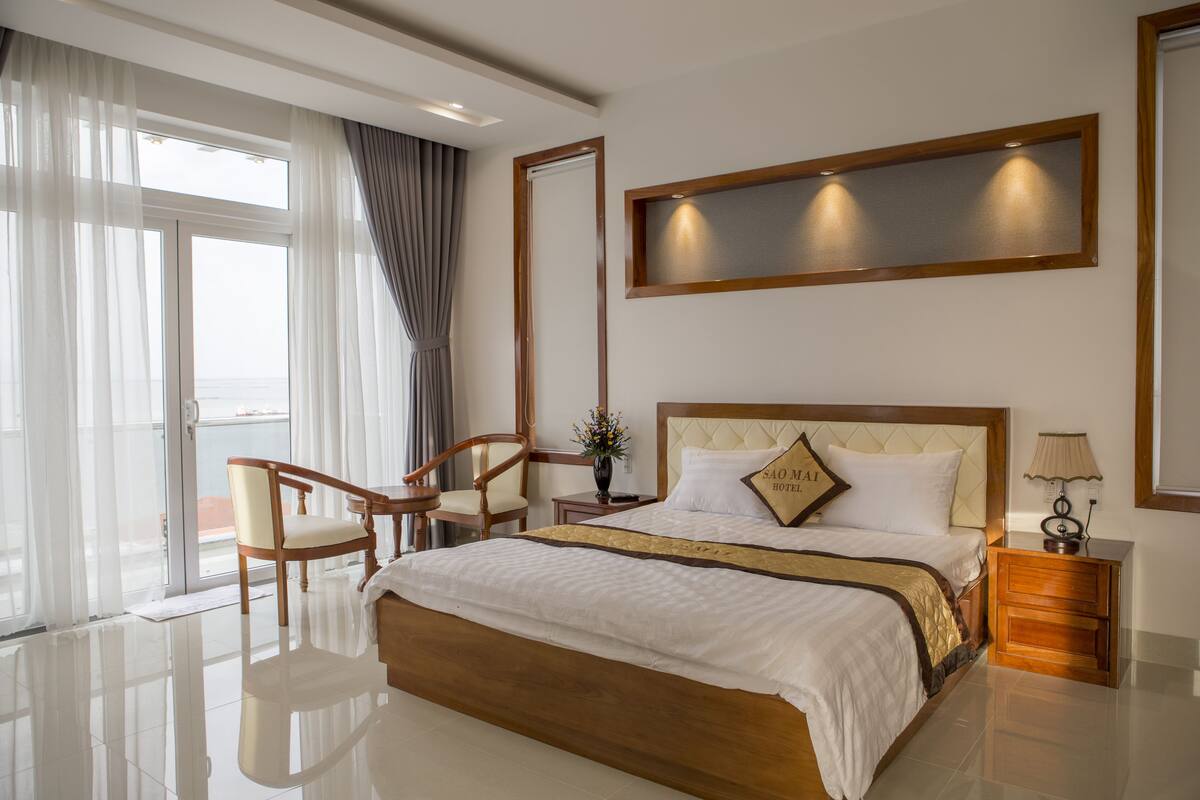 A hotel room with a large bed, a wooden nightstand, a chair, and a window with curtains.