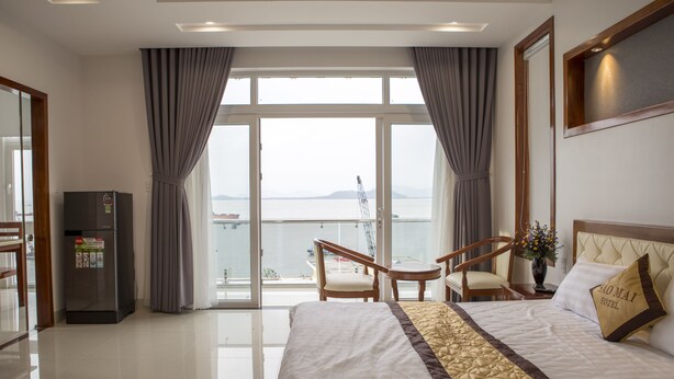 A hotel room with a large bed, a desk, a chair, a refrigerator, and a view of the sea.