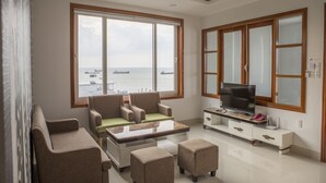 A modern living room with a sofa set, a coffee table, and a TV on a white cabinet. Large windows offer a view of the sea and boats.