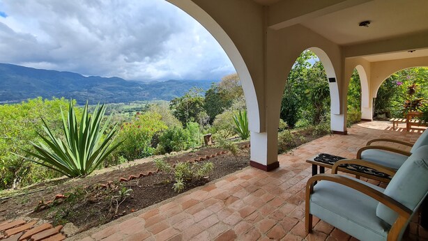 A covered outdoor seating area with brick flooring, white arches, and a view of a mountainous landscape.