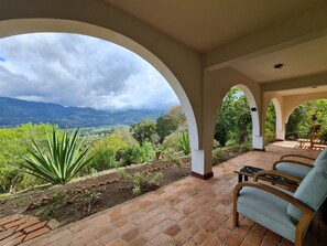 A covered outdoor seating area with brick flooring, white arches, and a view of a mountainous landscape. - Villas Valle Bello Ujarras (Santiago)