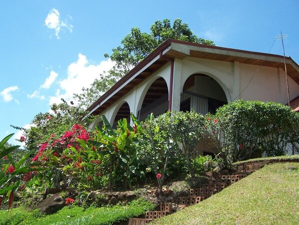 A house with a covered porch, surrounded by lush greenery and flowering plants. - Villas Valle Bello Ujarras (Santiago)