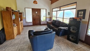 A living room with a wooden ceiling, a black leather sofa, a blue armchair, a wooden cabinet, and a large window with a view of the outdoors.