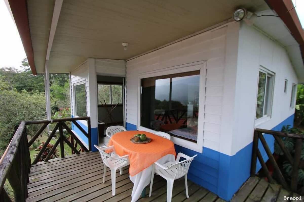 A covered outdoor seating area with a table and chairs, a window, and a view of the surrounding greenery.