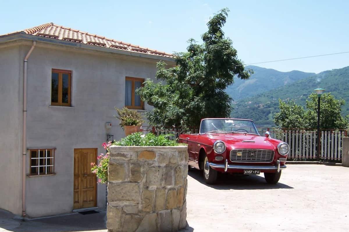 Une voiture rouge d’époque est garée devant une maison ornée d’un pilier en pierre et de plantes en pot.
