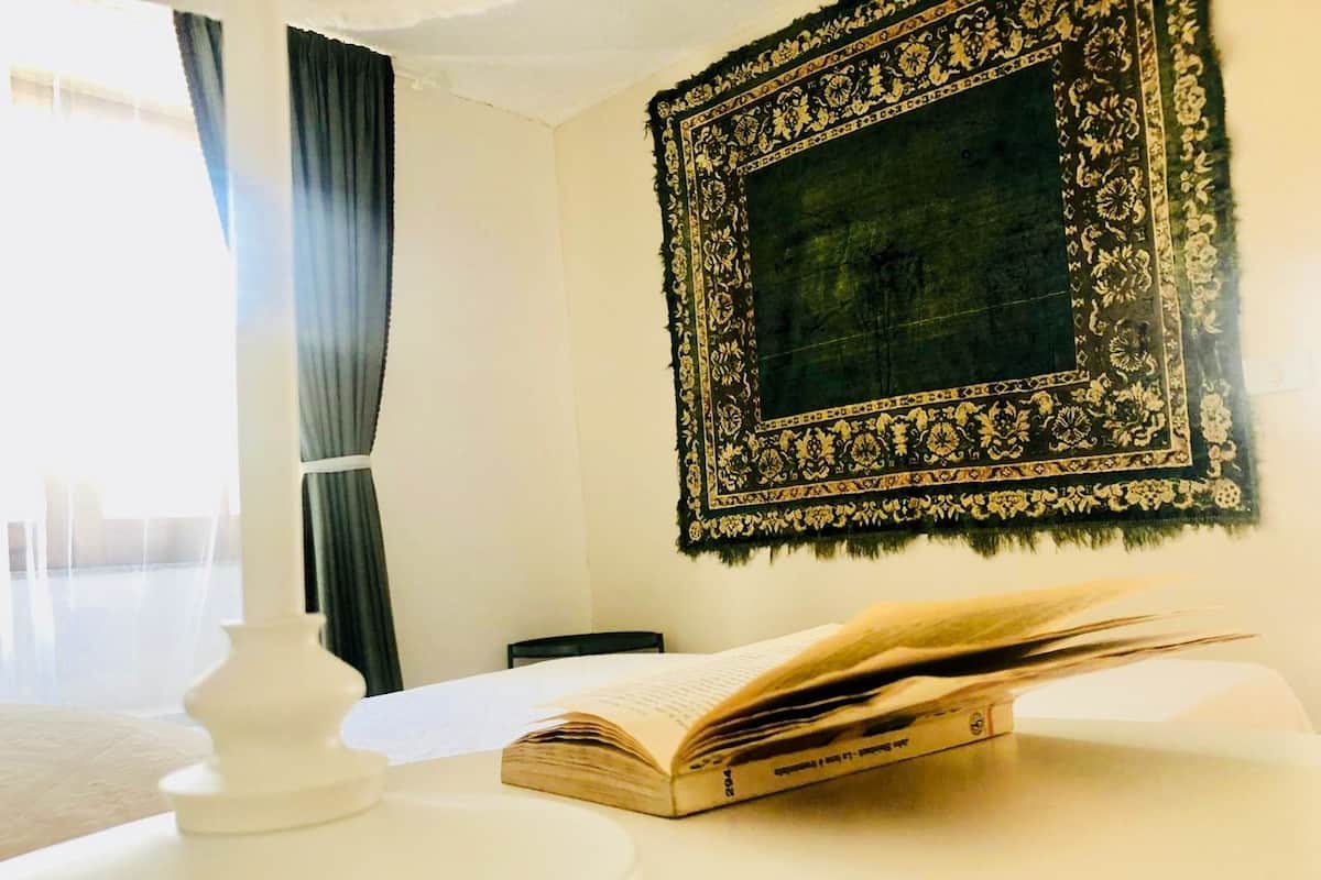 Une pièce avec une grande fenêtre, une table blanche sur laquelle repose un livre, une lampe blanche et une tapisserie à motifs accrochée au mur.