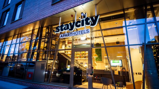 Een Staycity apartment hotel met een glazen gevel, interieurverlichting en een zichtbare trap naar de ingang.