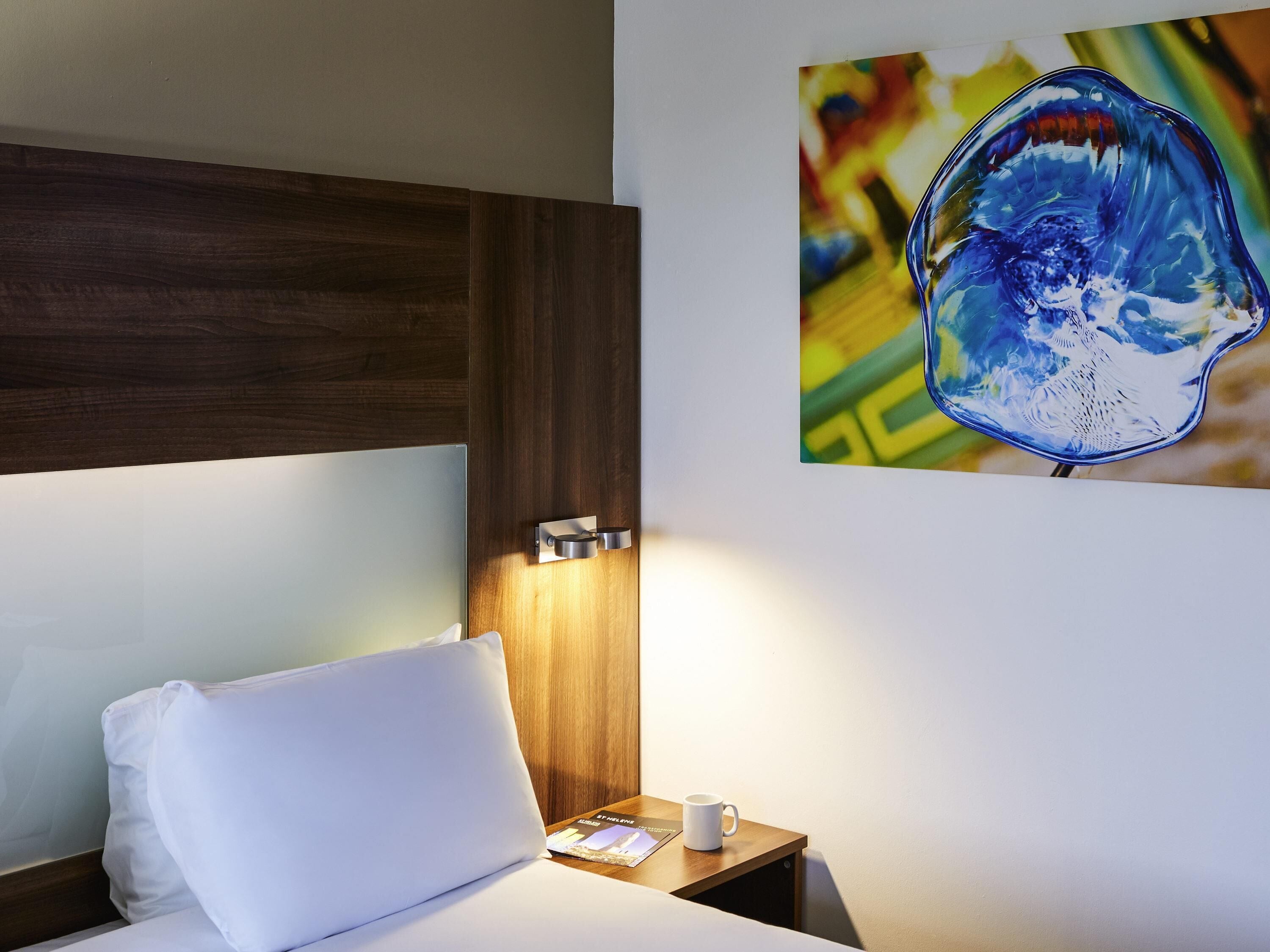 A hotel room with a bed, a nightstand, a white pillow, a white mug, and a wall-mounted light fixture.