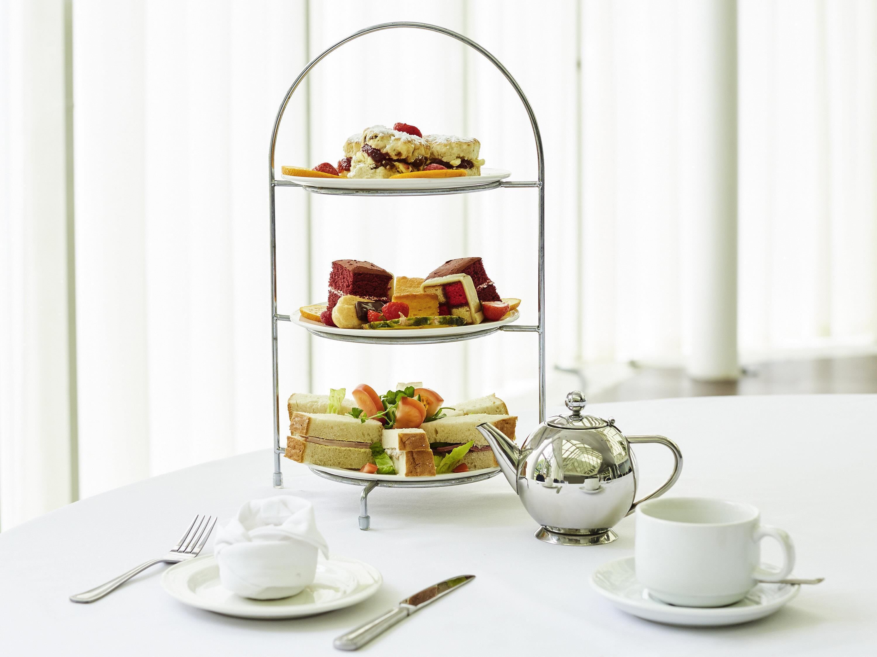 A three-tiered dessert stand with various pastries and sandwiches, a silver teapot, and a cup and saucer on a white table.