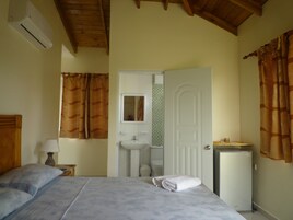 Un dormitorio con cama, una mesita de noche con lámpara, un refrigerador pequeño, un baño con lavamanos y espejo, y una puerta que da a otra habitación.
