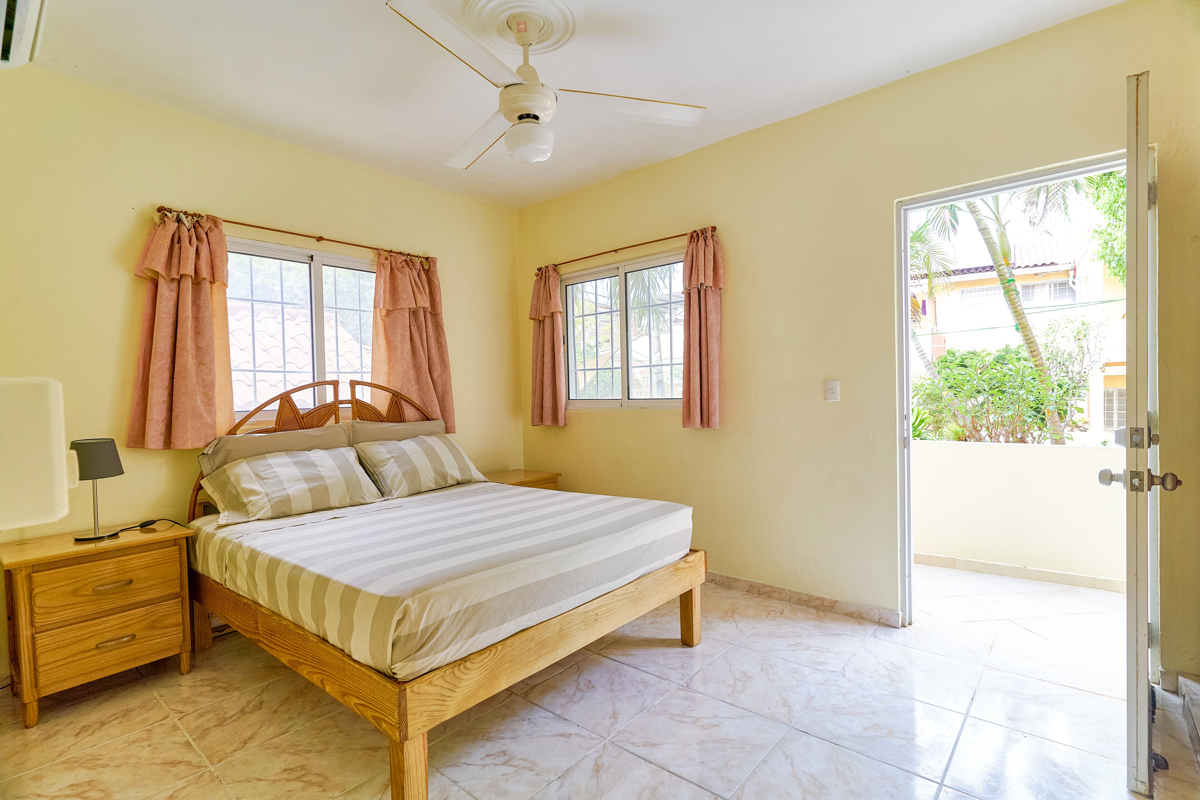 A bedroom with a bed, nightstand, and a ceiling fan. There are windows with curtains, and a door leading outside.