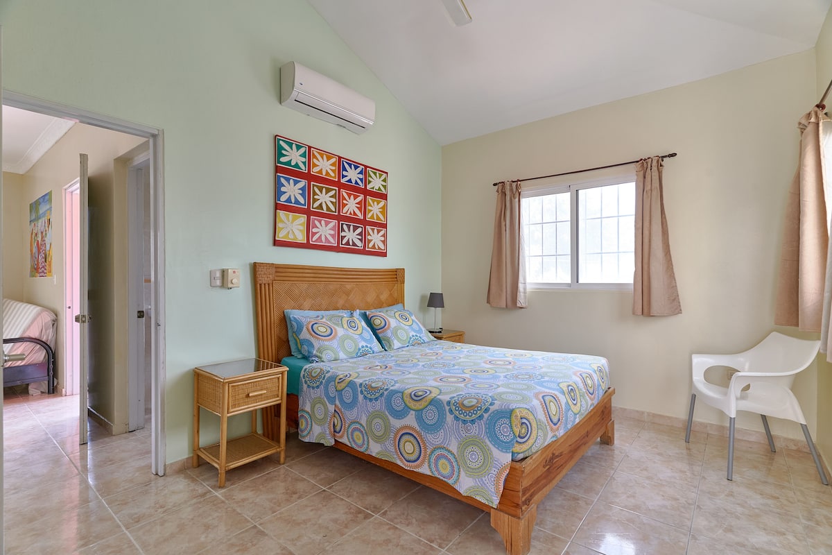 A bedroom with a bed, a bedside table, a chair, a window with curtains, and a wall-mounted air conditioner.
