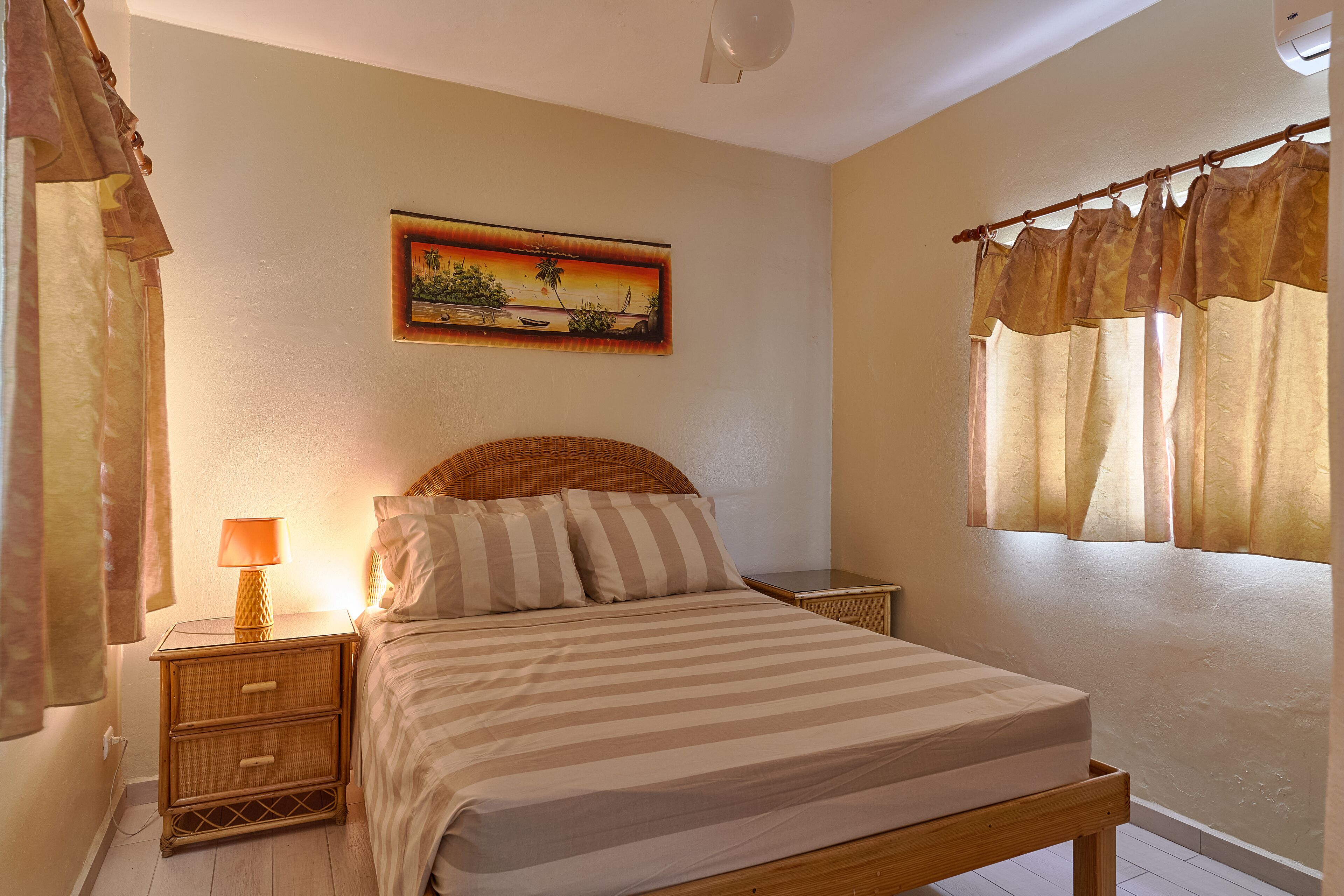 Une chambre à coucher avec un lit, une table de chevet, une lampe, un tableau au mur et une fenêtre avec des rideaux.