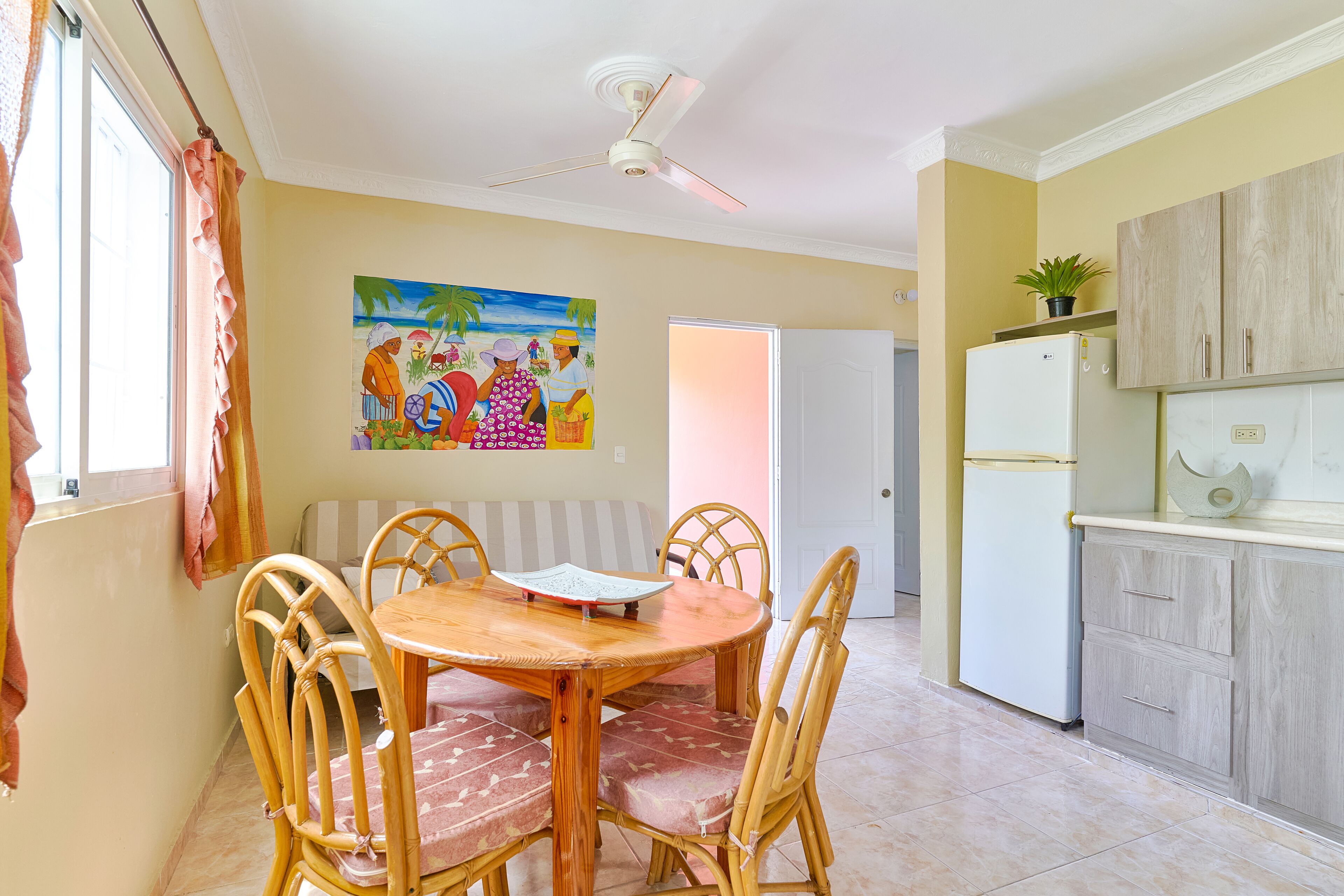 Un comedor con una mesa redonda de madera y sillas, un ventilador de techo, un cuadro en la pared y una cocina con armarios y un refrigerador.