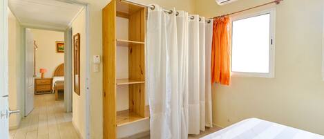A bedroom with a bed, a wooden wardrobe, a window with an air conditioner, and a shower area with a curtain.