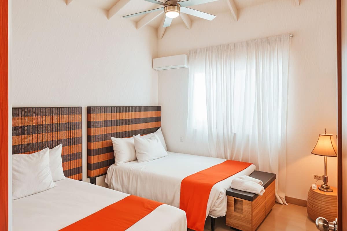 Ein Hotelzimmer mit zwei Betten, einem Deckenventilator, einer Klimaanlage, einem Nachttisch mit Lampe und einem Fenster mit Vorhängen.