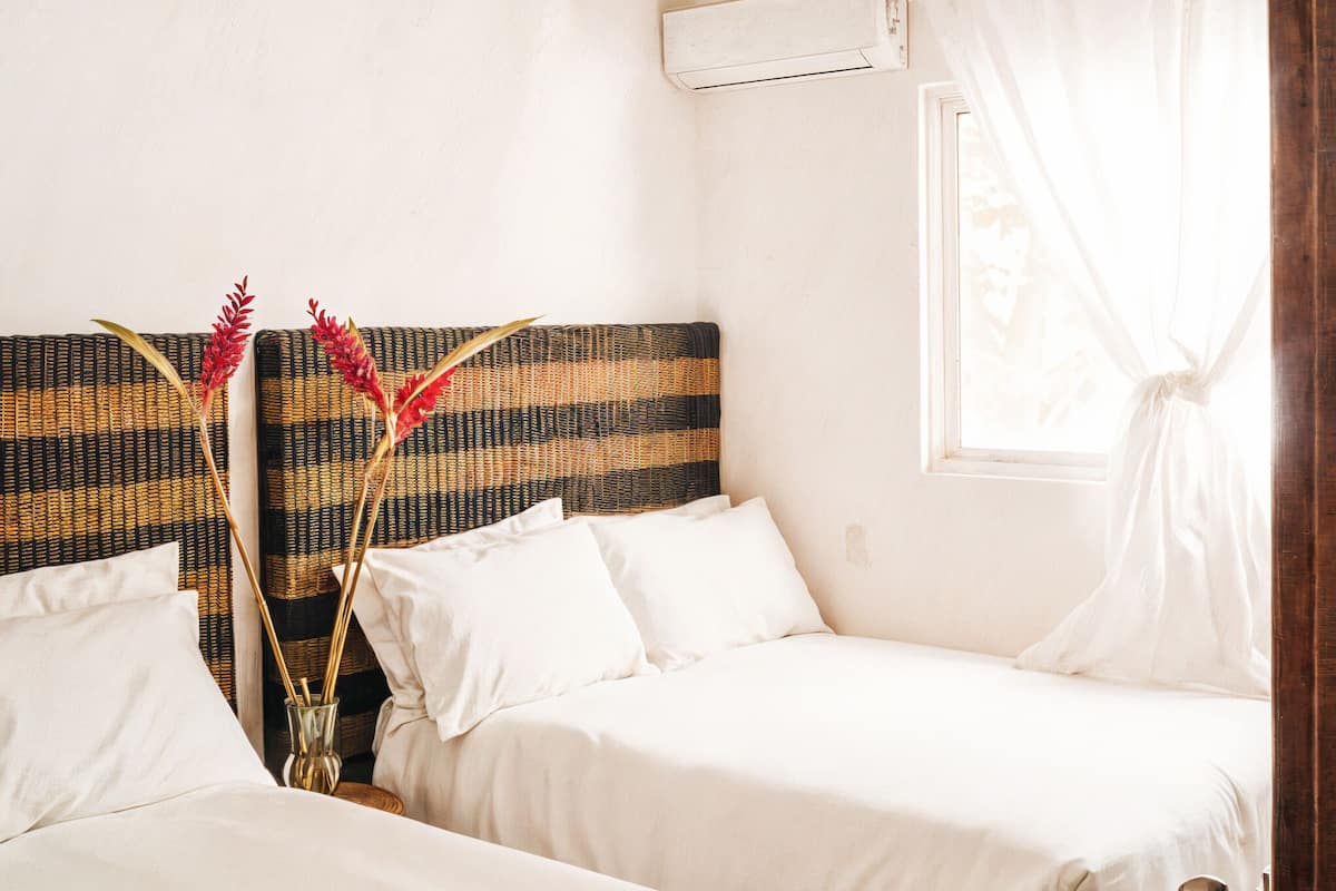 Ein Zimmer mit zwei Betten, einem gewobenen Kopfteil, weißer Bettwäsche, einem Fenster mit durchsichtigen Vorhängen und einer Wand-Klimaanlage.
