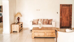 Una sala de estar con una mesa de centro de madera, un sofá blanco con cojines estampados, una mesita de noche de madera con una lámpara y una puerta de madera.