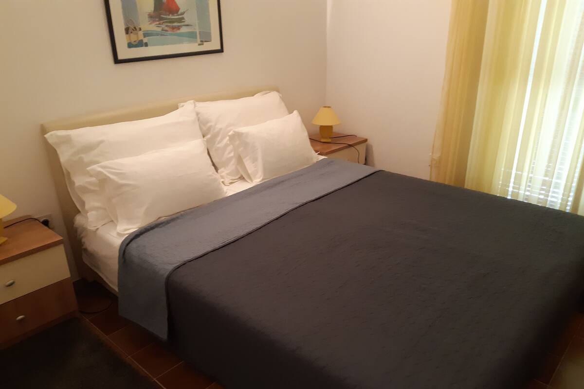 A neatly made bed with white pillows and a grey bedspread in a room with a small wooden nightstand, a lamp, and a framed picture on the wall.