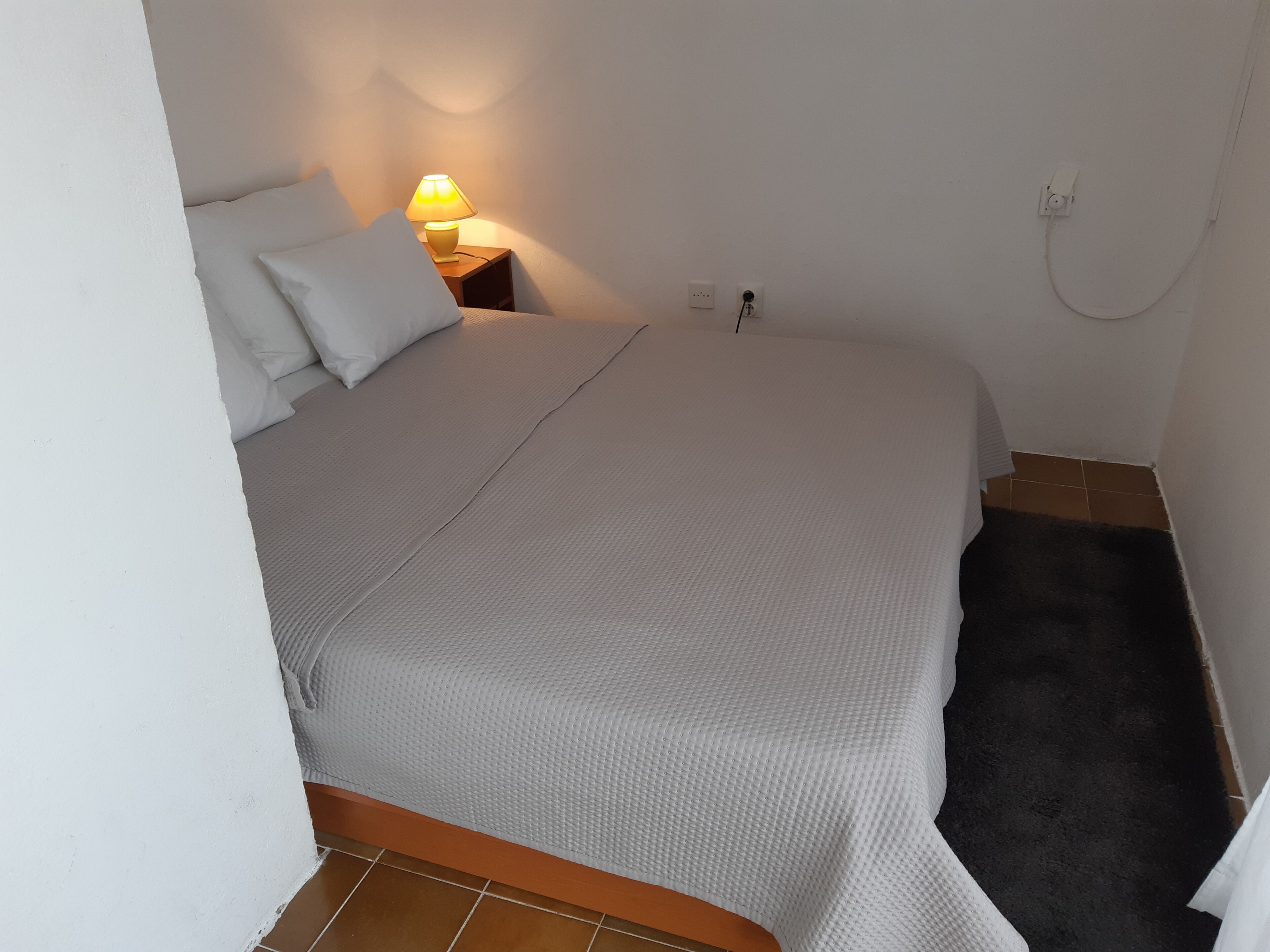 A neatly made bed with a white duvet, a bedside lamp, and a power outlet on the wall.