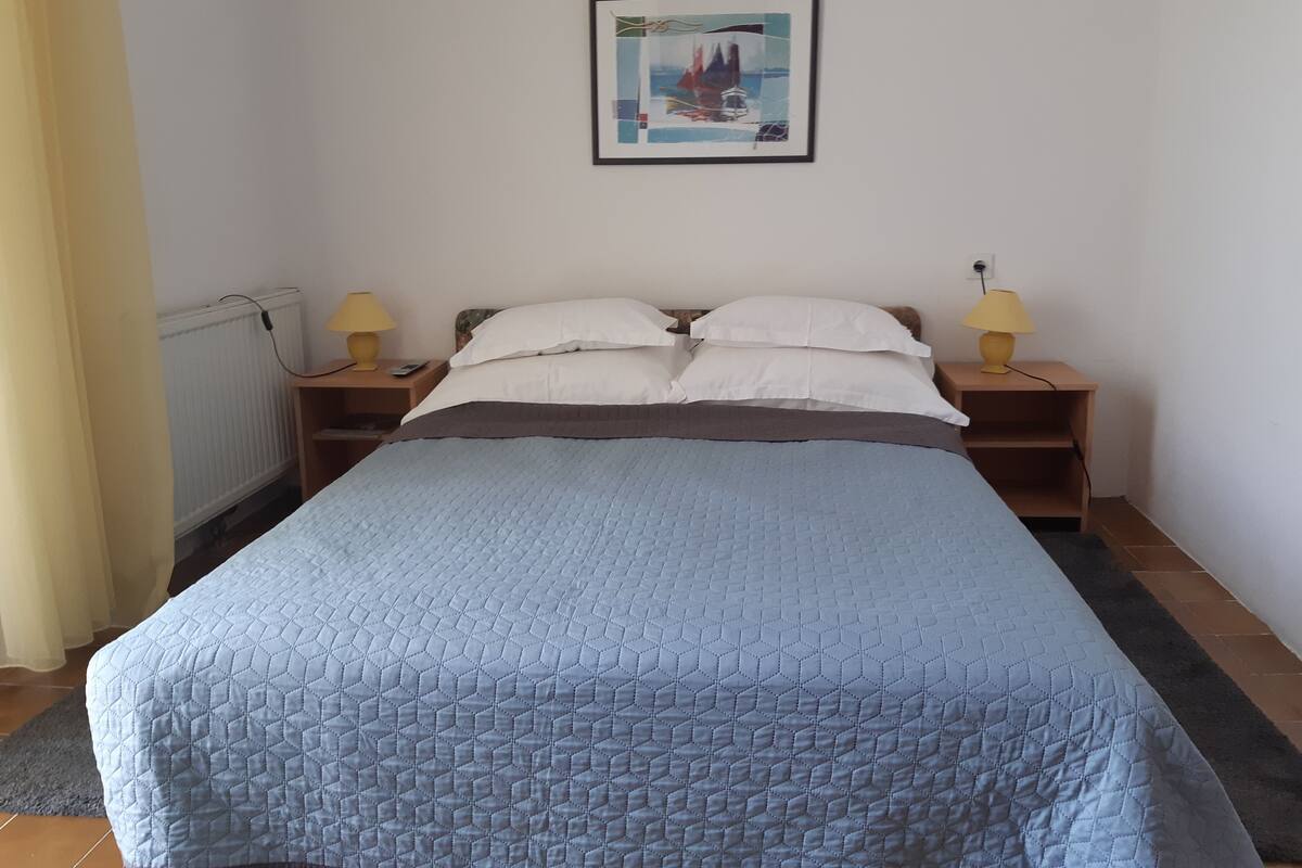 A bedroom with a bed, two bedside tables, two lamps, a framed picture on the wall, and a radiator.