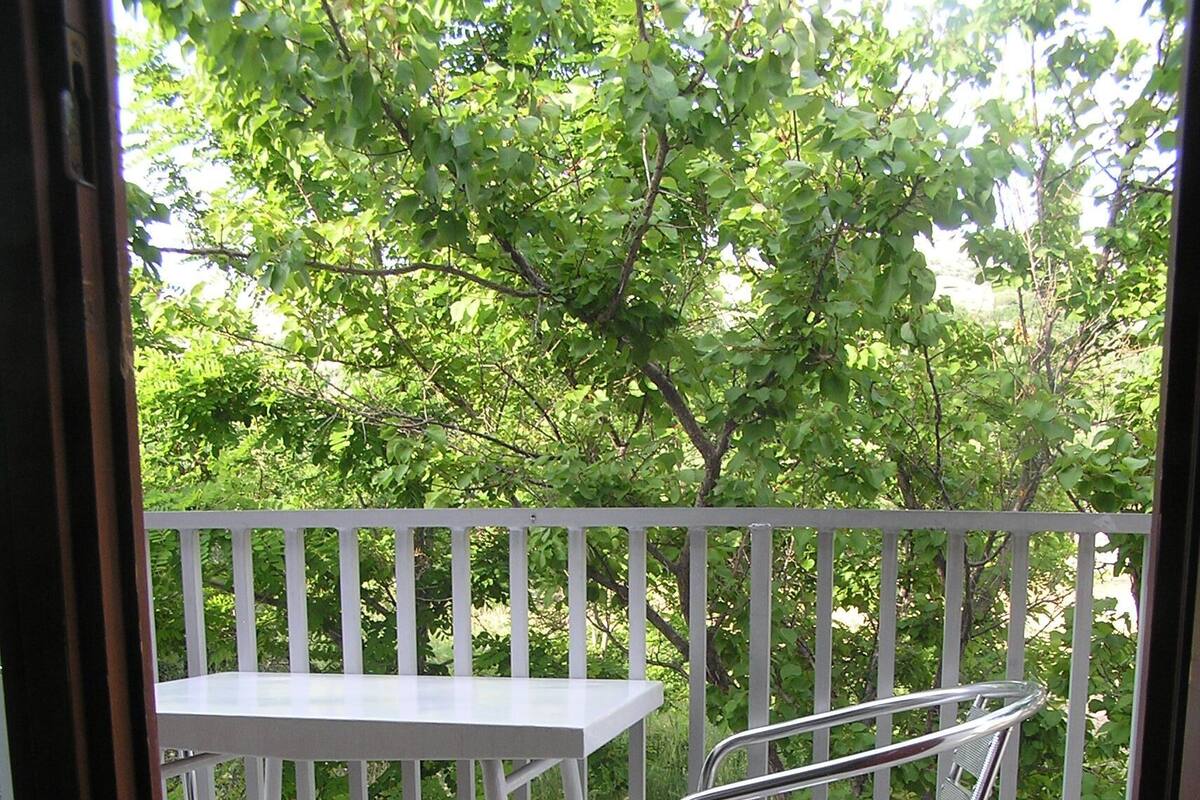 A balcony with a white table and metal chair, surrounded by a white railing and greenery.