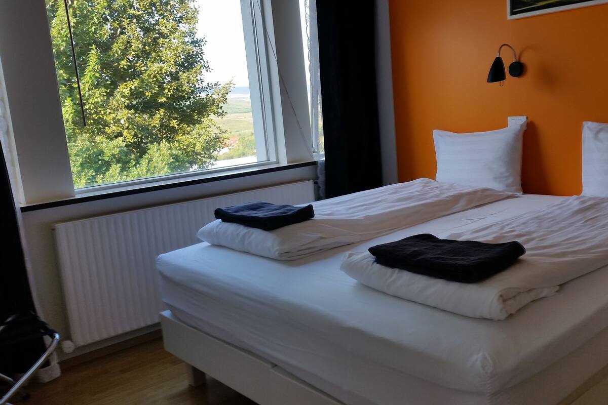 Cama bem arrumada com lençóis brancos e duas almofadas, janela com vista para o verde, e parede com luminária preta.